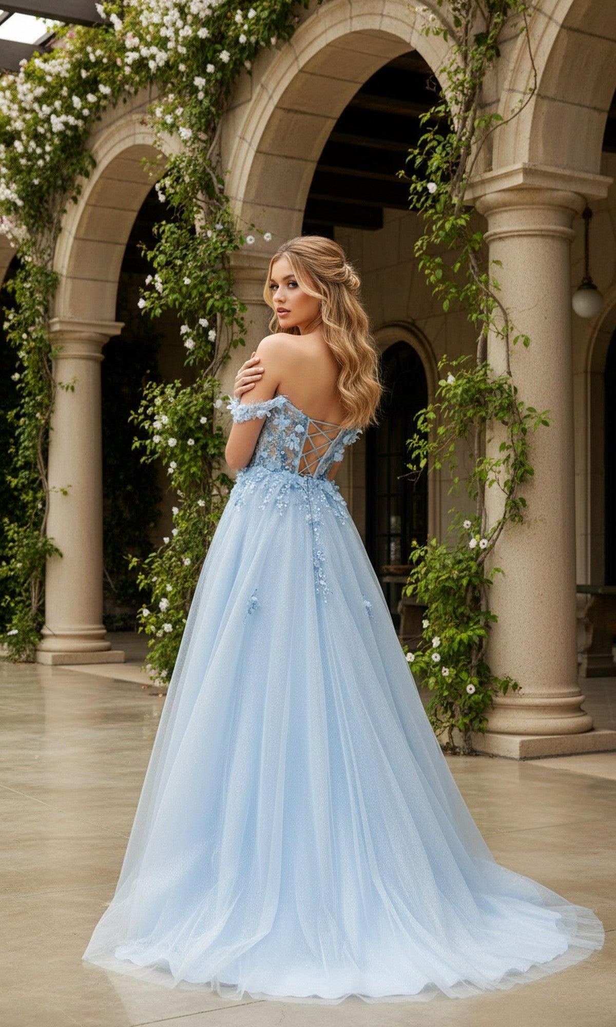 A woman in the Long Prom Dress: Cinderella Couture 8403J, a light blue off-the-shoulder gown with 3D flower details, stands before an arched stone building draped in greenery.